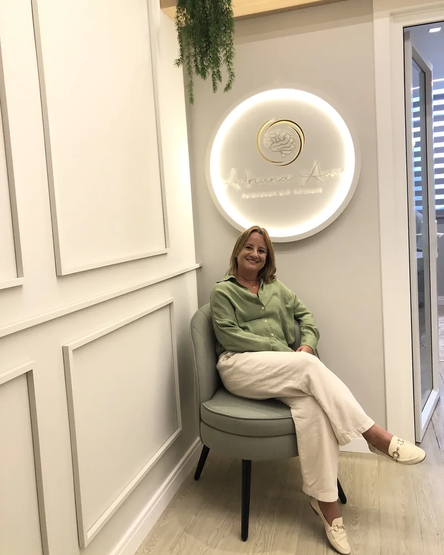 Adriana Assis sentada numa poltrona na recepção do consultório, ao lado do painel iluminado com o seu logo