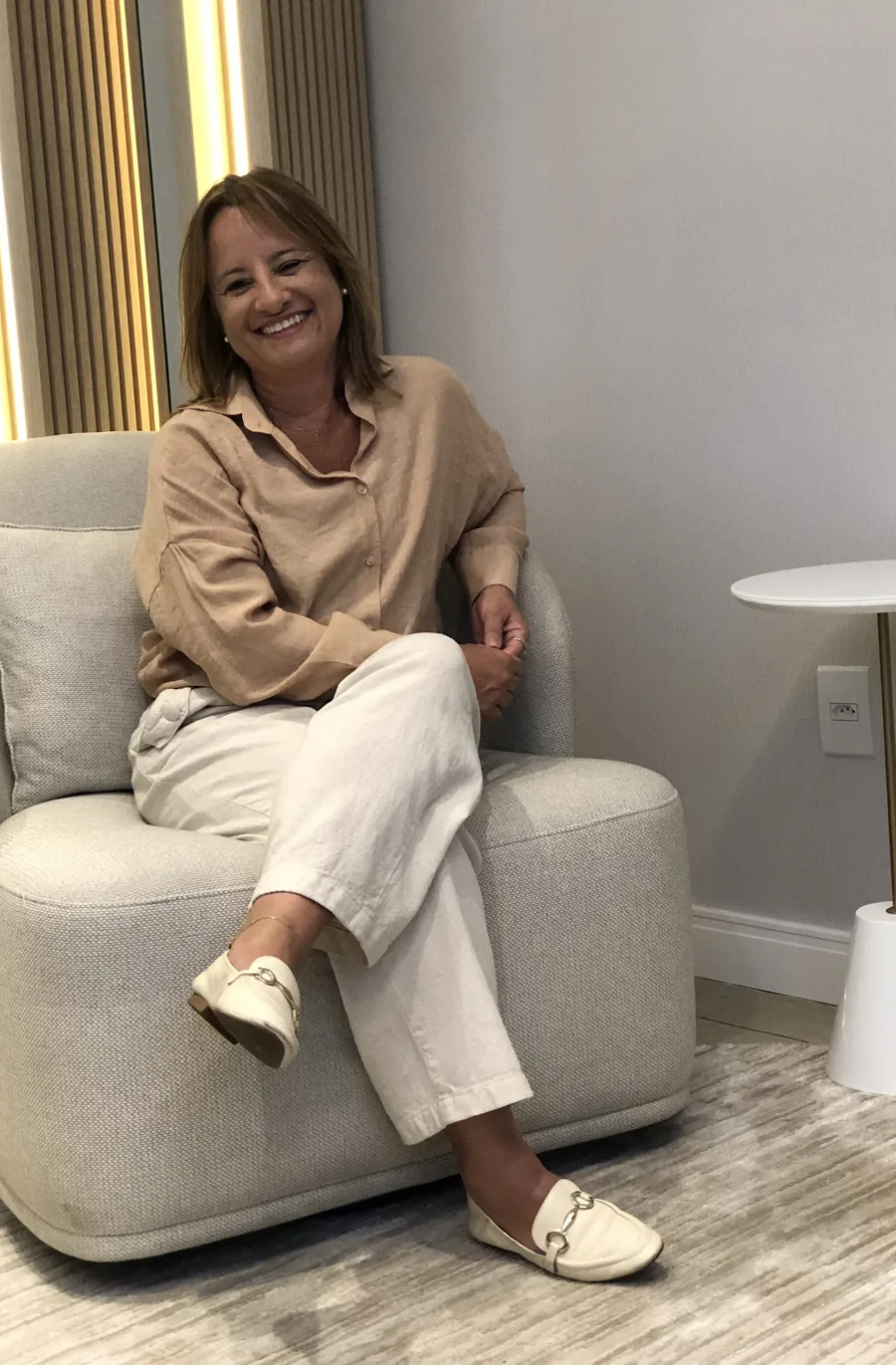 Adriana Assis rindo, de camisa bege e calça clara, sentada de pernas cruzadas numa poltrona do consultório